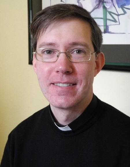 Ein Mann mit Brille und schwarzem Priesterhemd lächelt im Innenbereich in die Kamera.