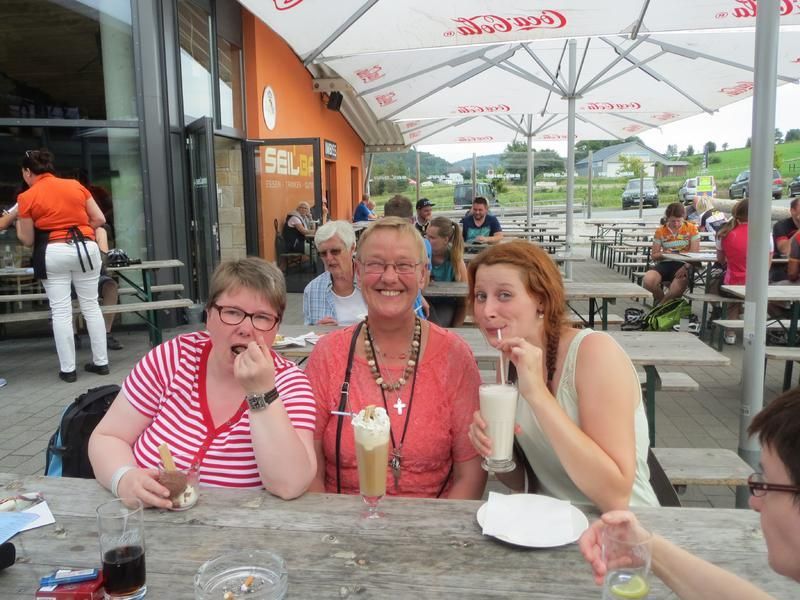 Drei Frauen genießen Getränke in einem Straßencafé. Andere sitzen in der Nähe; darüber ein orangefarbenes Gebäude und ein Sonnenschirm.