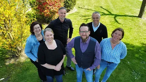 Eine Gruppe von sechs lächelnden Personen posiert gemeinsam in einem sonnigen Garten.