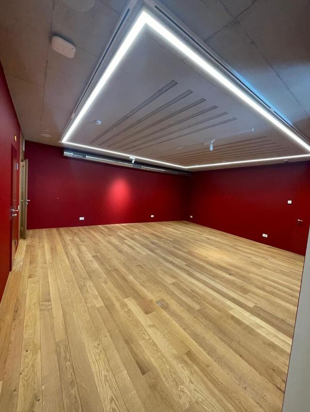Leerer Raum mit roten Wänden, hellem Holzboden und eingelassener Deckenbeleuchtung.