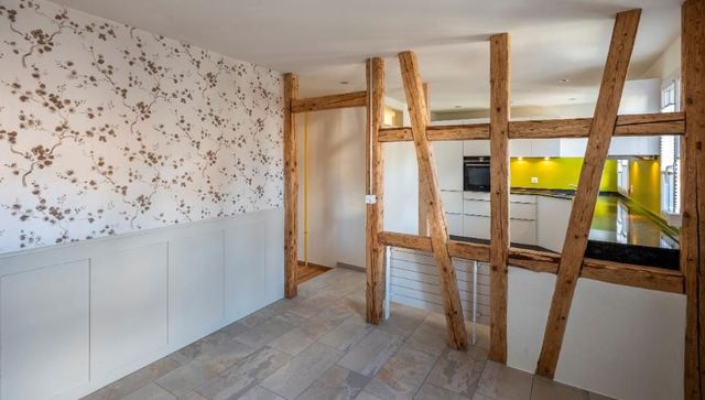 Innenansicht eines modernen Raumes mit freiliegenden Holzbalken, weissen Schränken und einer Küche mit gelbem Fliesenspiegel.