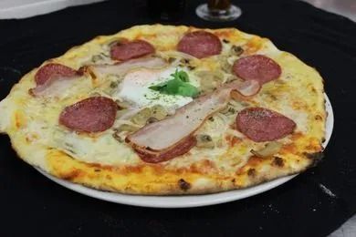 Une pizza avec du bacon, du salami et un œuf sur une assiette blanche sur une table.