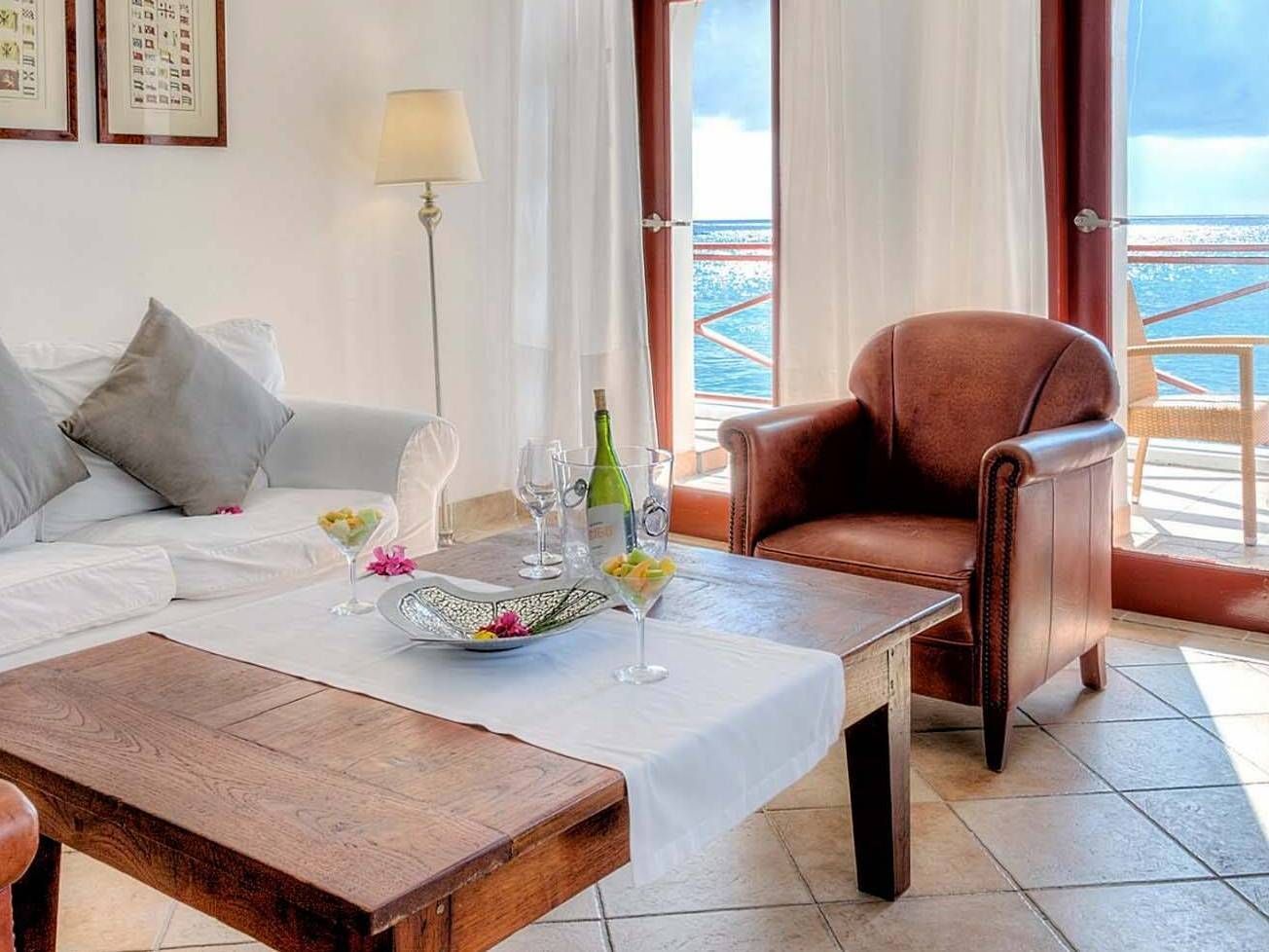 Wohnzimmer mit Meerblick, weißes Sofa, brauner Ledersessel, Holztisch mit Getränken.