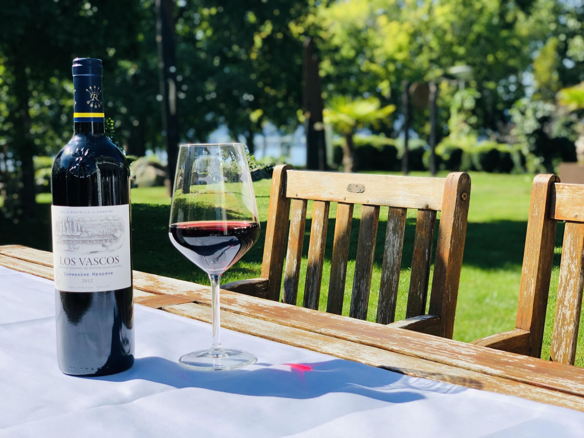 Eine Flasche Rotwein und ein mit Wein gefülltes Glas stehen auf einem Tisch im Freien, im Hintergrund ist ein Garten zu sehen.