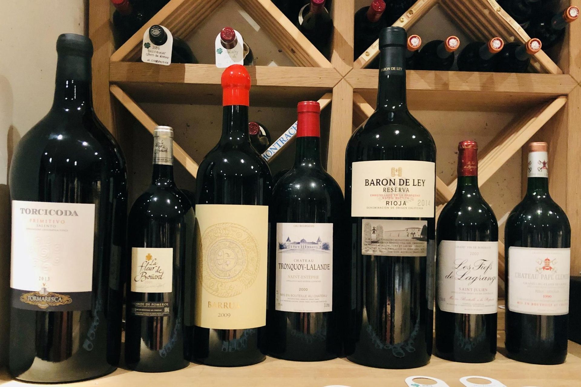 Weinflaschen verschiedener Größen auf einem Holzregal, einige mit roten Verschlüssen.