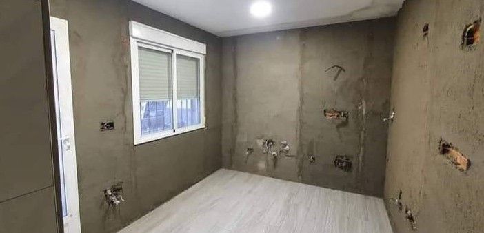 Una habitación vacía y sin terminar, con paredes de yeso gris, una ventana blanca, suelo de madera clara y tuberías