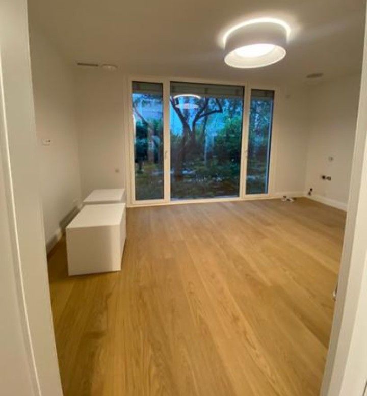 Una habitación vacía con suelo de madera clara, dos bancos cúbicos blancos y una gran puerta de cristal