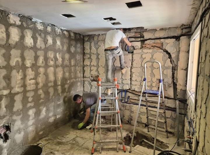 Dos trabajadores renuevan una habitación con paredes de piedra vista; uno está subido a una escalera cerca del techo