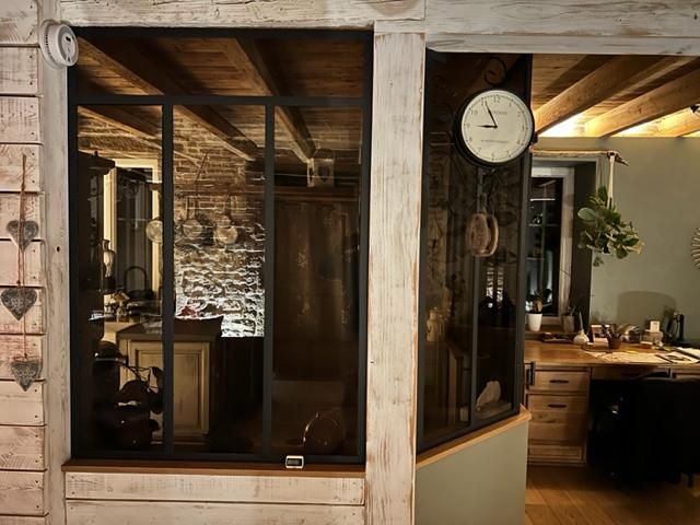 Un intérieur rustique avec une cloison vitrée à cadre noir séparant une cuisine aux murs en pierre d'un espace de travail avec un bureau.