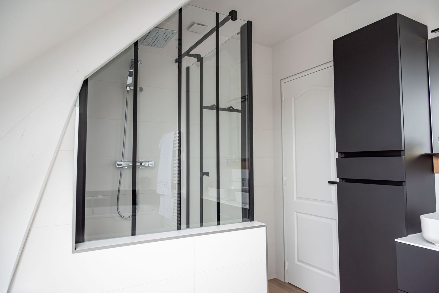 Une cabine de douche en verre à cadre noir dans une salle de bains aux murs blancs inclinés et un meuble sombre.