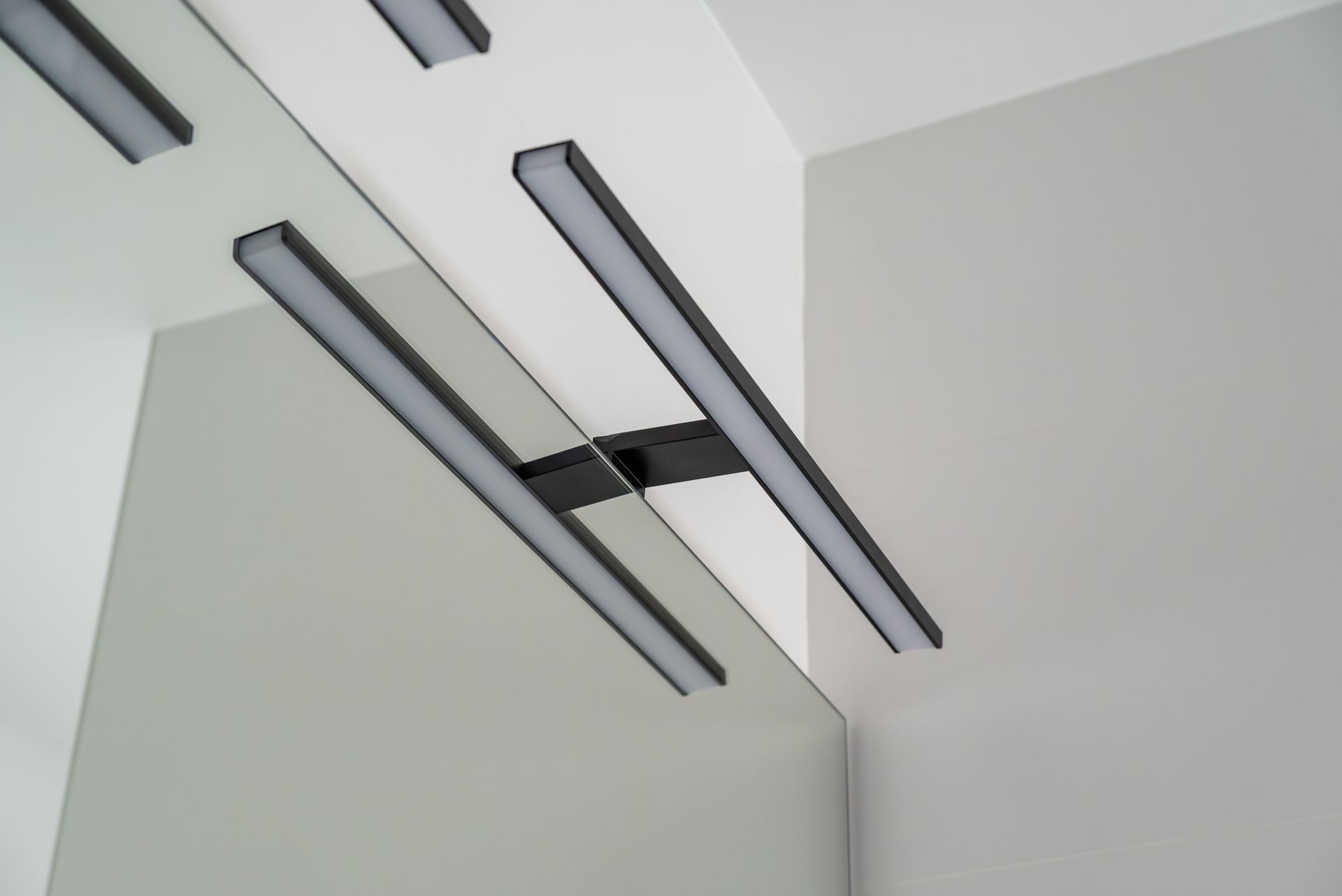Un luminaire linéaire LED noir à double barre, fixé dans un coin de mur au-dessus d'un miroir, réfléchissant la lumière.