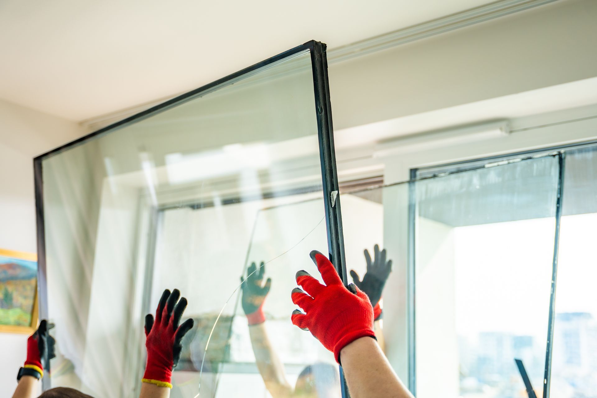 Des ouvriers portant des gants de sécurité rouges soulèvent une grande vitre pour la mettre dans son cadre lors de travaux de rénovation d'une maison.