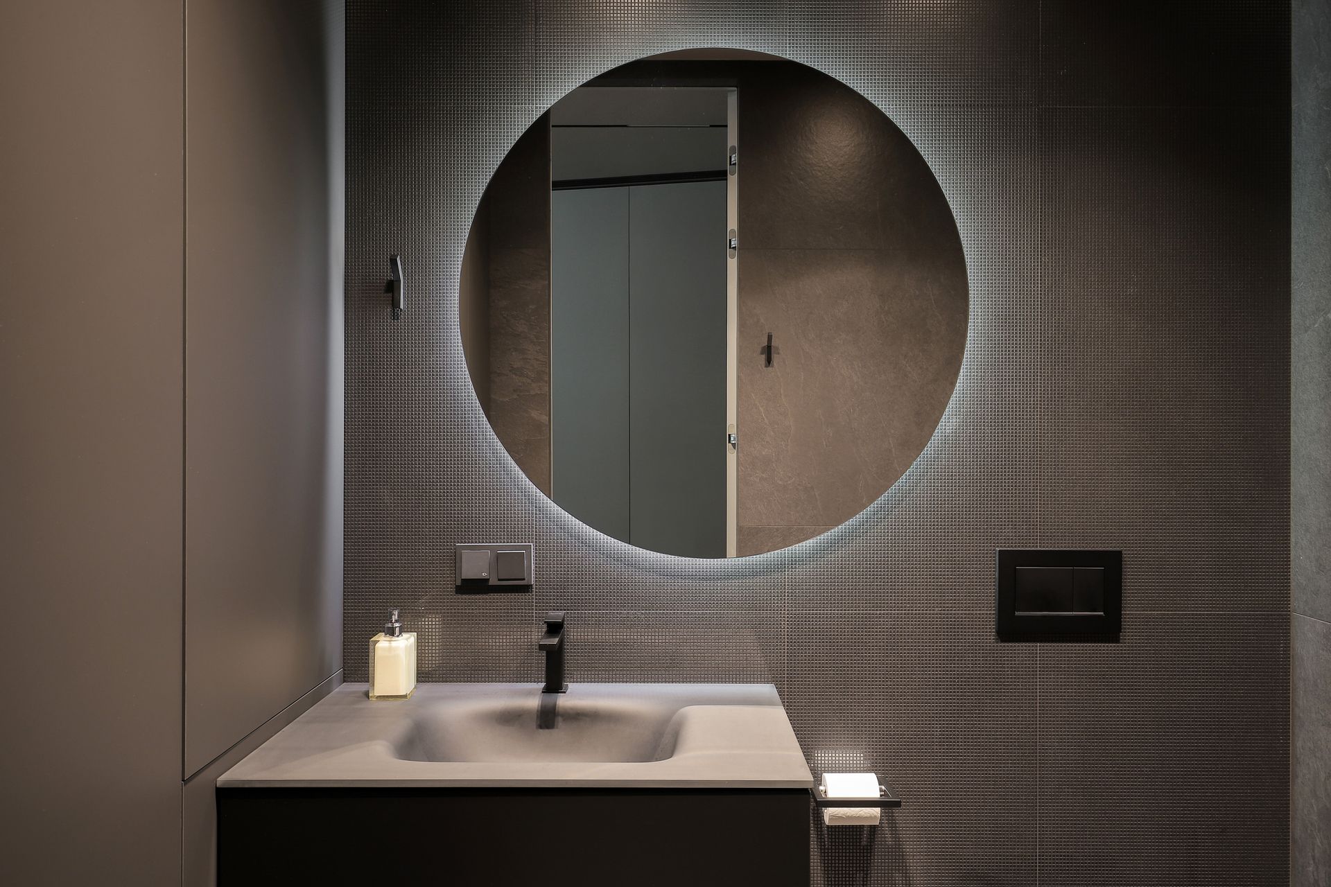Meuble-lavabo moderne avec un lavabo carré, un robinet noir mat et un miroir rond rétroéclairé sur un mur à motif quadrillé foncé.