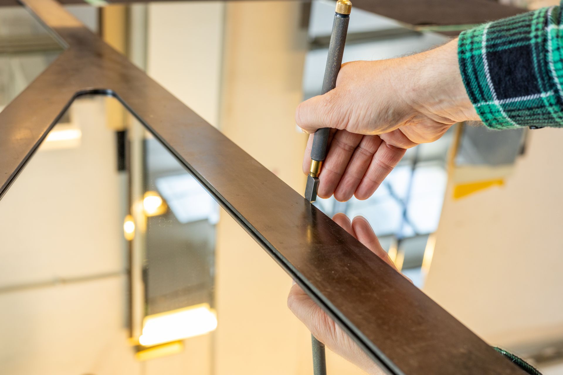 Une main tenant un coupe-verre portatif trace une ligne sur la surface d'un miroir le long d'un guide métallique.