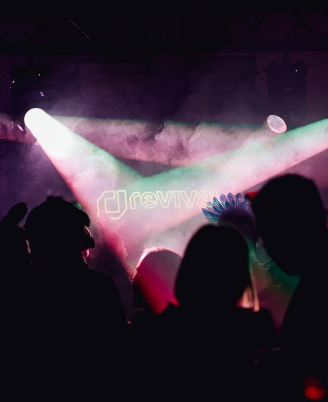 Siluetas del público en un concierto, con luces rosas y verdes en el escenario y un haz de luz de un foco