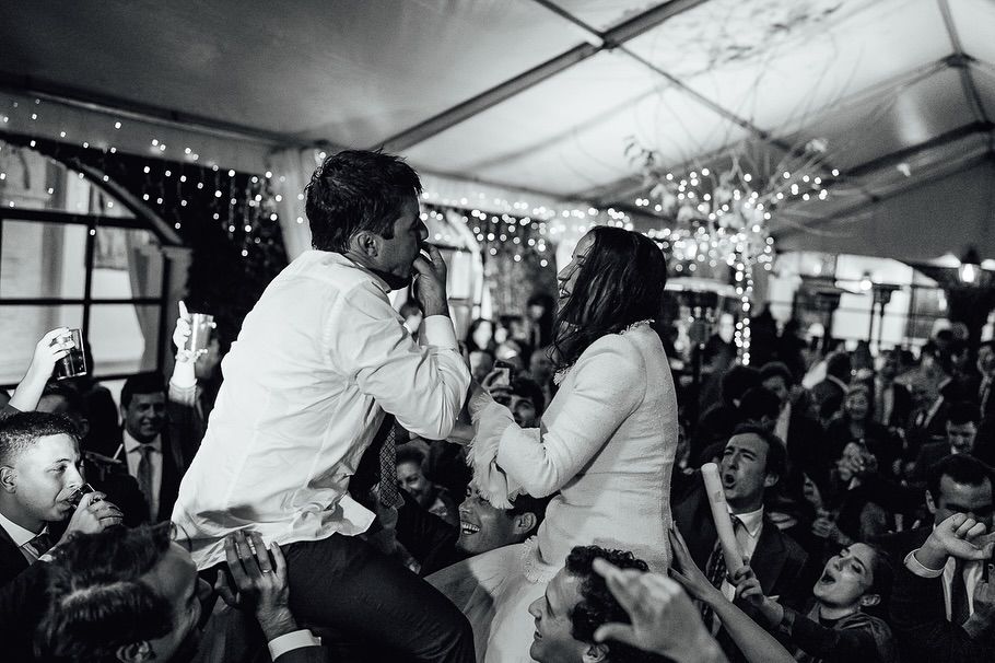 Escena en blanco y negro de una multitud con dos personas cantando en el escenario bajo una carpa, con guirnaldas de luces brillando detrás de ellas
