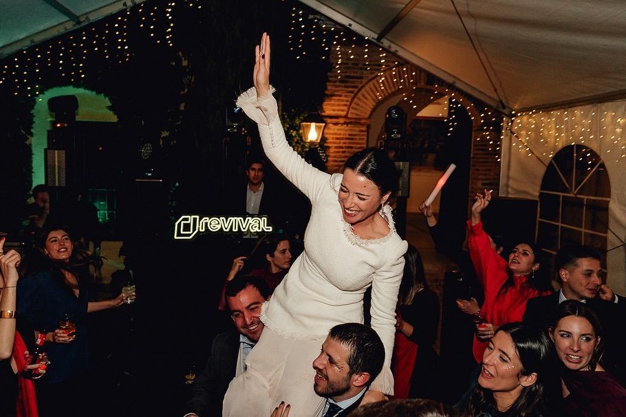Gente bailando en una fiesta animada en un recinto cerrado; una mujer vestida de blanco, subida a hombros de alguien, levanta la mano por encima de la cabeza