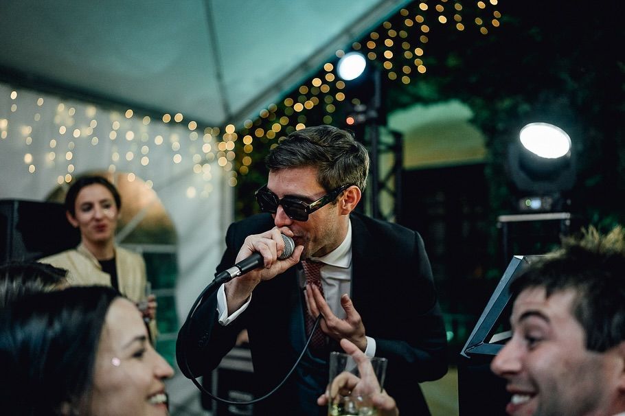 Un cantante actúa en una animada fiesta en un espacio cerrado, con un micrófono en la mano, mientras los invitados lo miran y sonríen bajo guirnaldas de luces