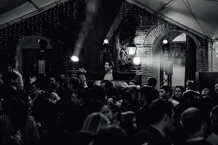 Una multitud reunida frente a un escenario de concierto interior tenuemente iluminado, bajo arcos y luces colgantes