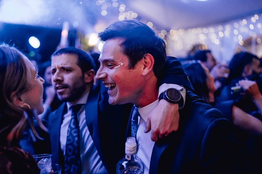 Personas sonrientes y conversando en una fiesta iluminada de azul; una de ellas tiene el brazo sobre los hombros de otra.