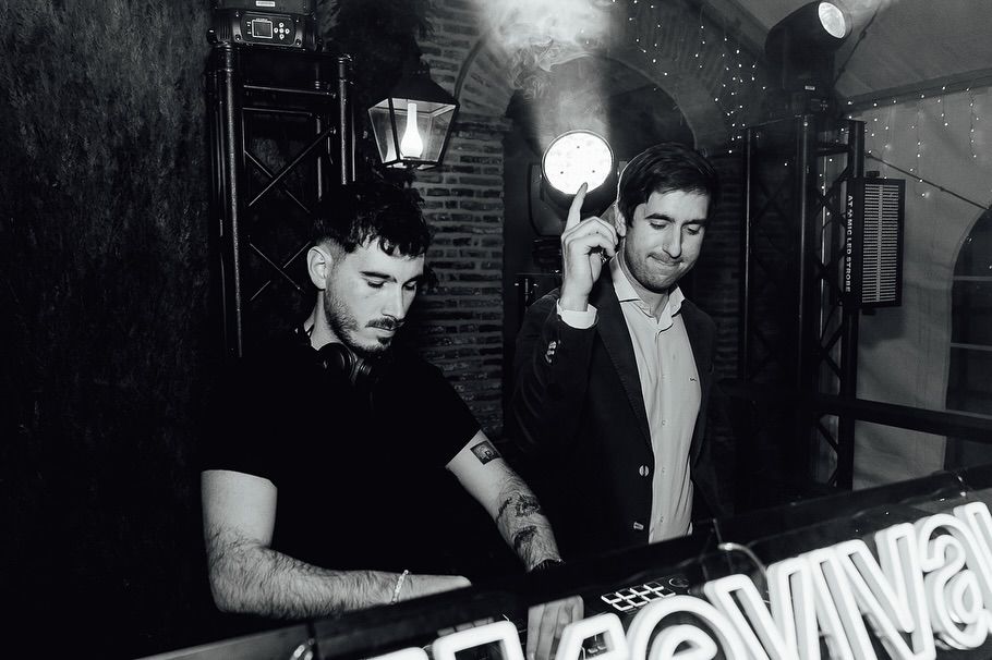 Dos DJ en la cabina de una discoteca, uno levantando una luz brillante, con un letrero de neón delante