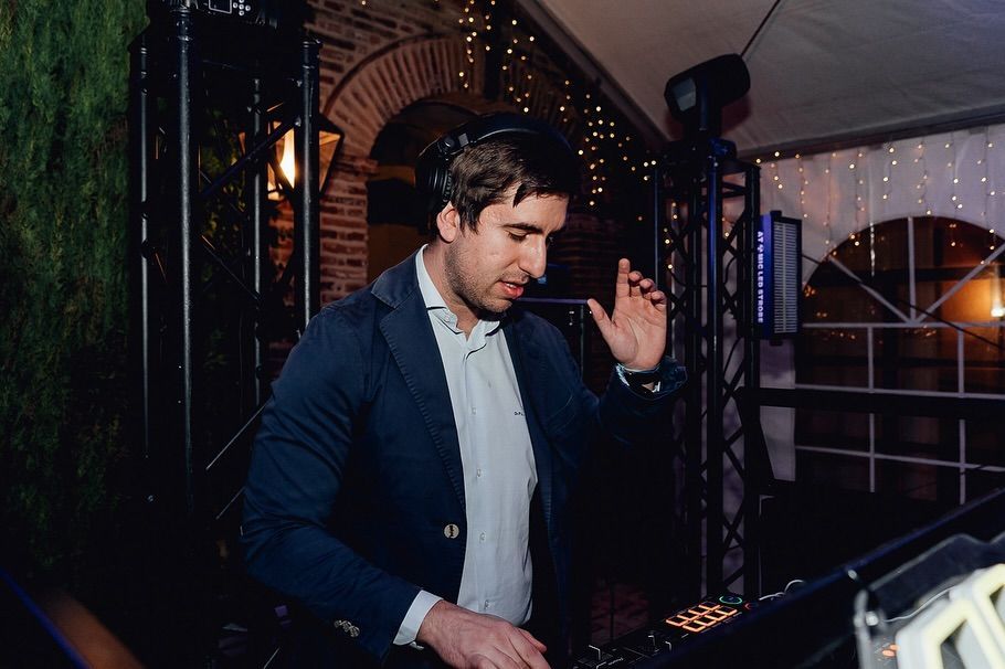 Un DJ con una chaqueta azul oscuro mezclando música en un evento nocturno con luces cálidas y un fondo en forma de arco