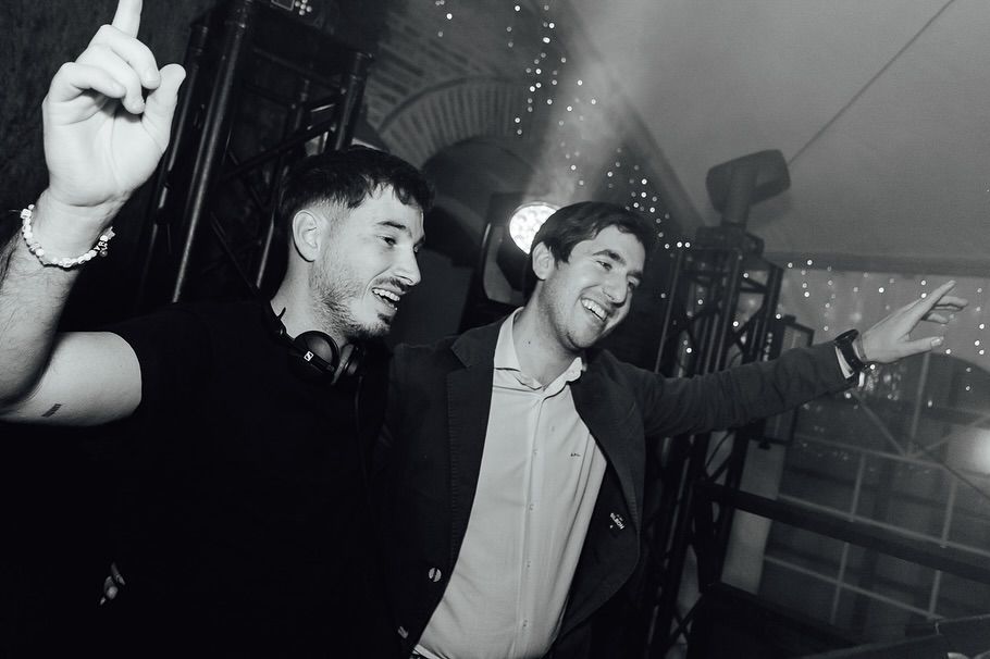 Dos hombres sonríen y bailan en una discoteca, mientras uno levanta la mano bajo las luces del escenario.