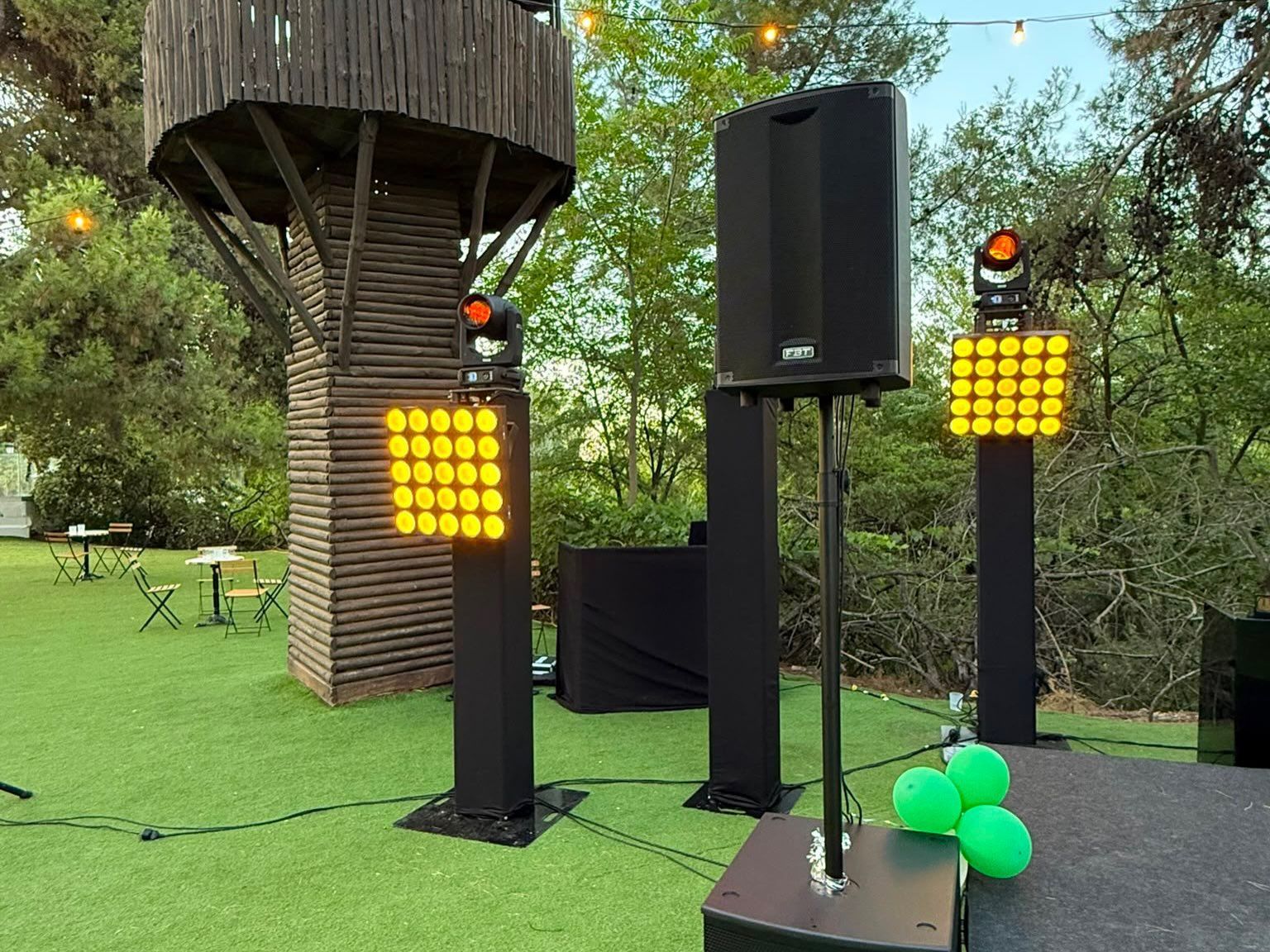 Escenario al aire libre con altavoces negros, luces amarillas y globos verdes sobre el césped, bajo los árboles