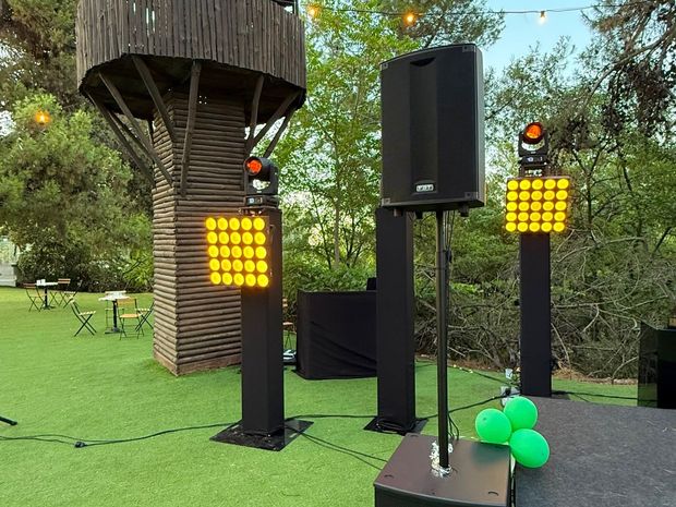 Escenario al aire libre con altavoces negros, luces amarillas y globos verdes sobre el césped, bajo los árboles