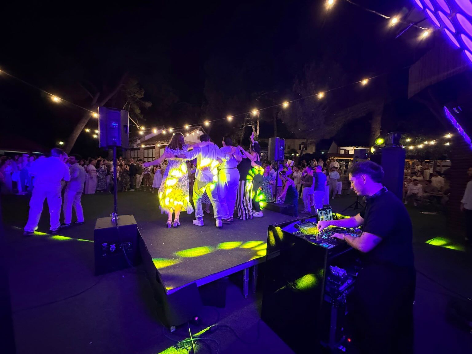 Personas bailando en un escenario al aire libre bajo luces moradas y amarillas, con un DJ en una mesa cerca.