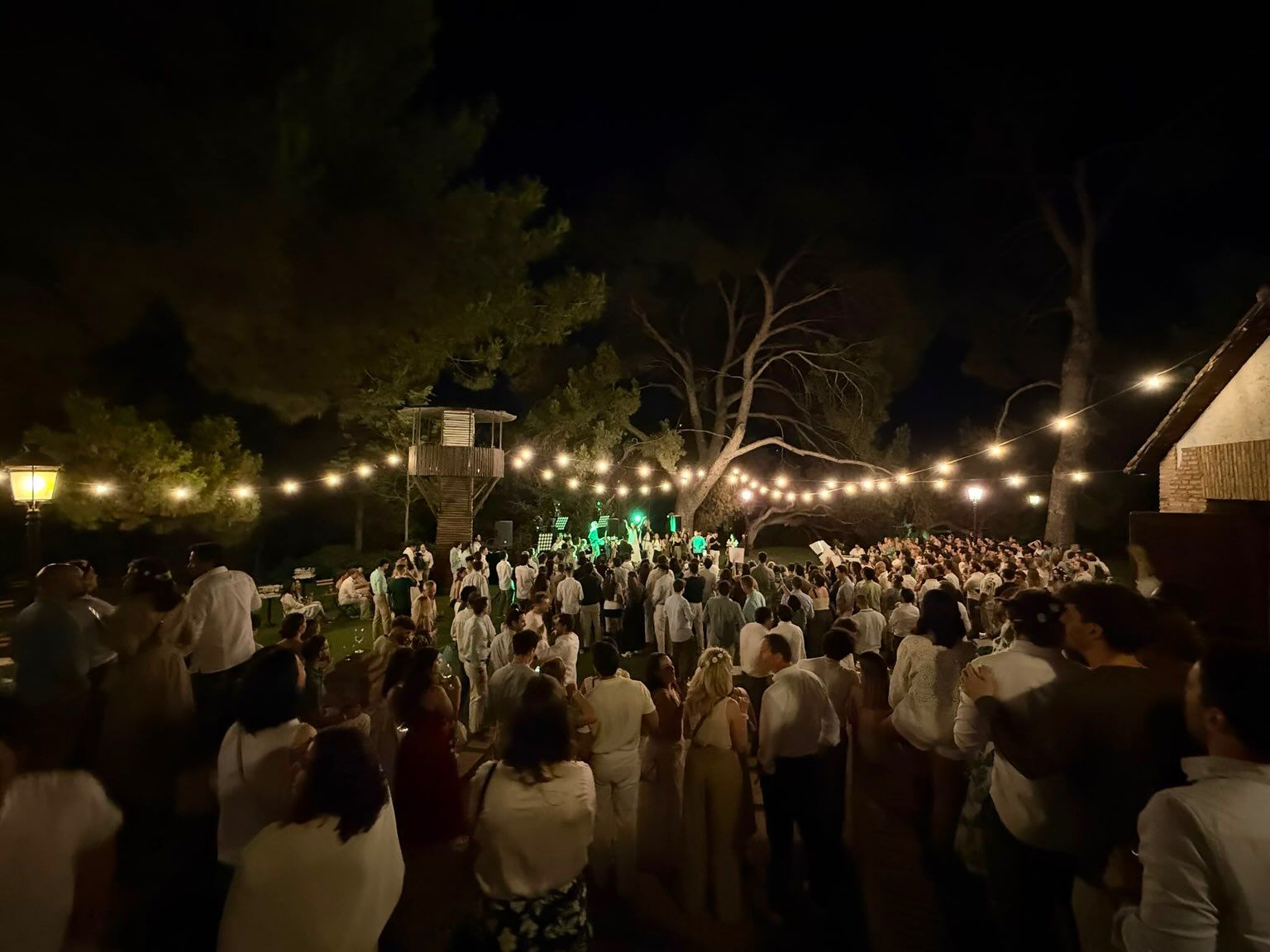 Aglomeración nocturna al aire libre bajo guirnaldas de luces, con la gente mirando hacia un escenario o altar iluminado
