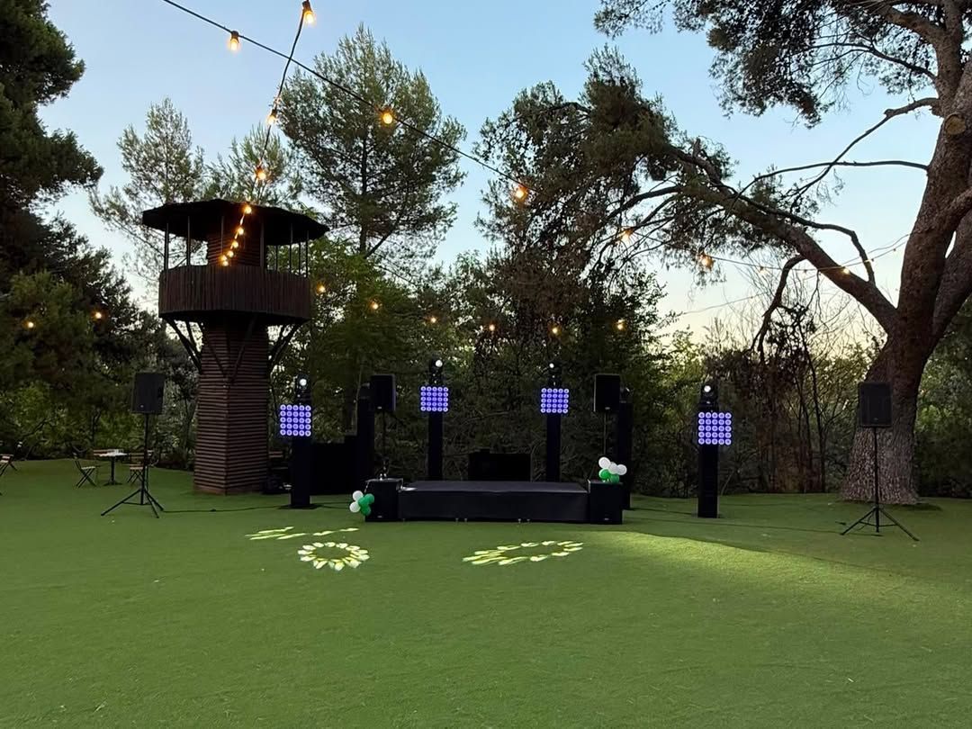 Escenario al aire libre al anochecer en un parque con césped, con postes iluminados, una plataforma elevada y árboles altos sobre él