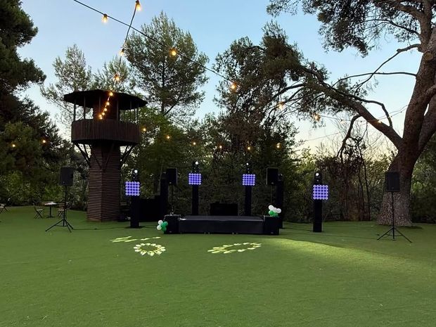 Escenario al aire libre al anochecer en un parque con césped, con postes iluminados, una plataforma elevada y árboles altos sobre él