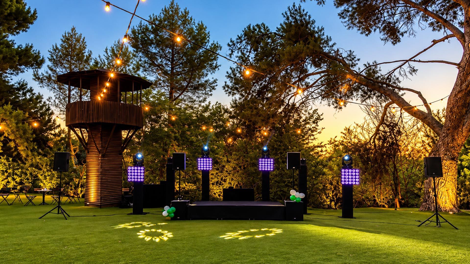 Escenario al aire libre en un jardín iluminado al anochecer, con guirnaldas de luces y farolillos morados bajo los árboles