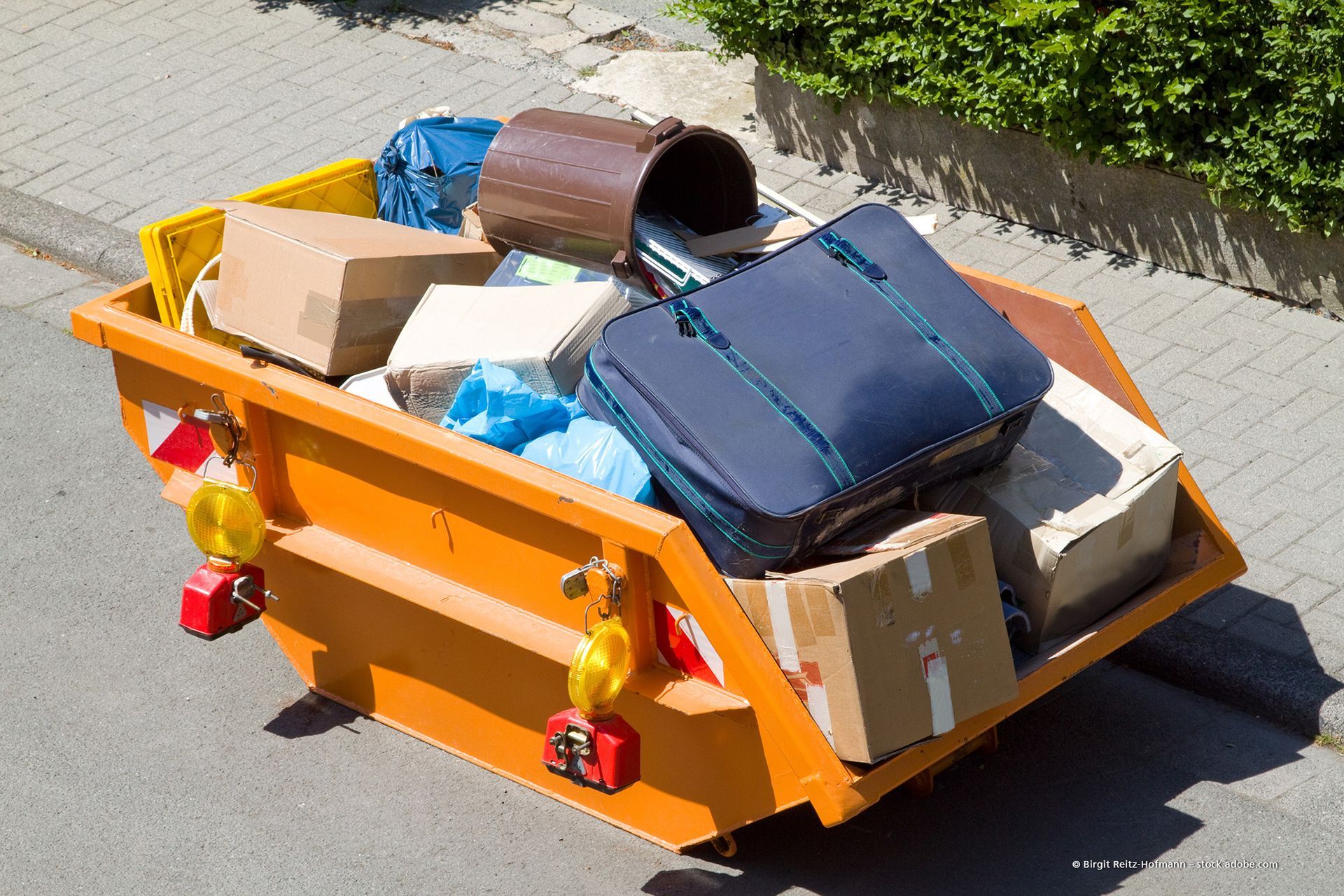 Ein grüner Müllcontainer, überquellend mit ausrangierten Möbeln, steht auf einer Rasenfläche in der Nähe einer Straße.