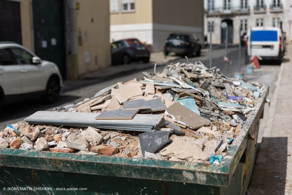 Bauschutt in einem Müllcontainer auf einer Straße in der Stadt; im Hintergrund Autos und Gebäude.