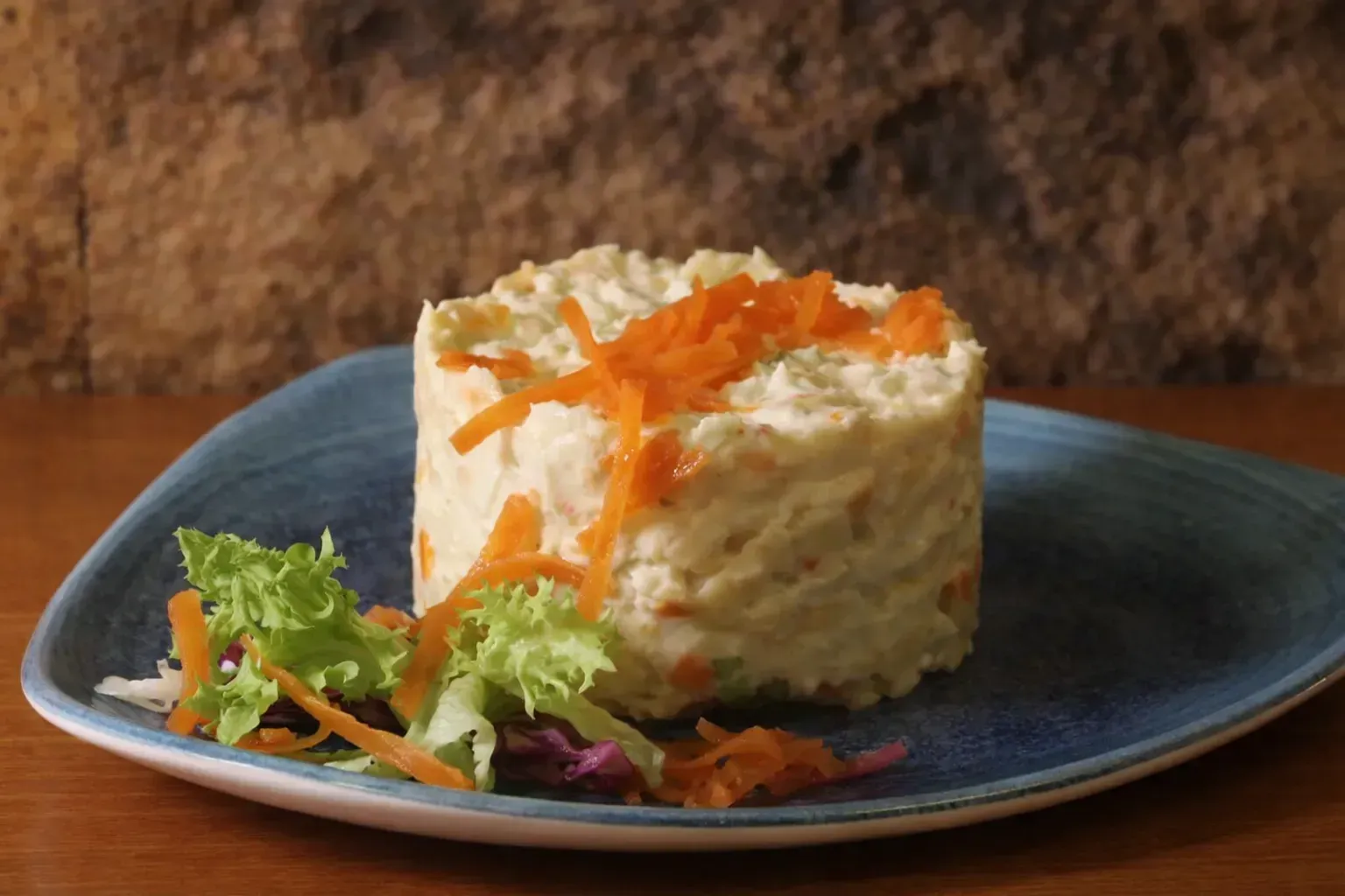 Una ensalada de papas cilíndrica con guarnición de zanahoria y una ensalada en un plato azul.