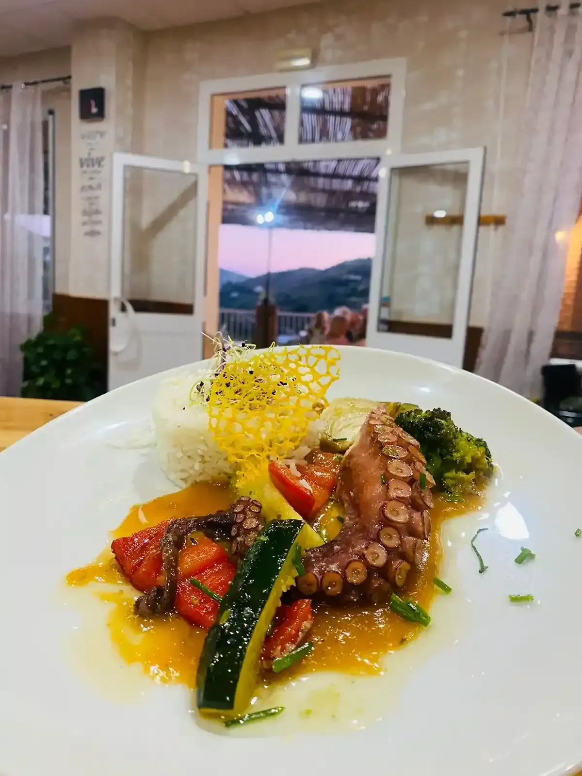 Plato de pulpo con arroz, verduras y salsa, ambiente de restaurante con vistas a la montaña.
