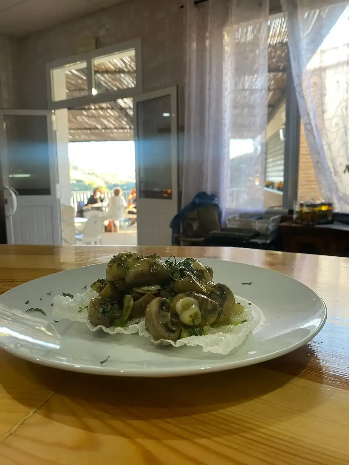 Plato de champiñones con hierbas sobre una mesa de madera, interior de cafetería con una vista al aire libre al fondo.