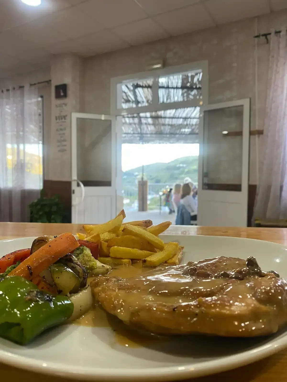 Plato con carne en salsa, patatas fritas y verduras a la plancha, con puerta que da a una vista panorámica.