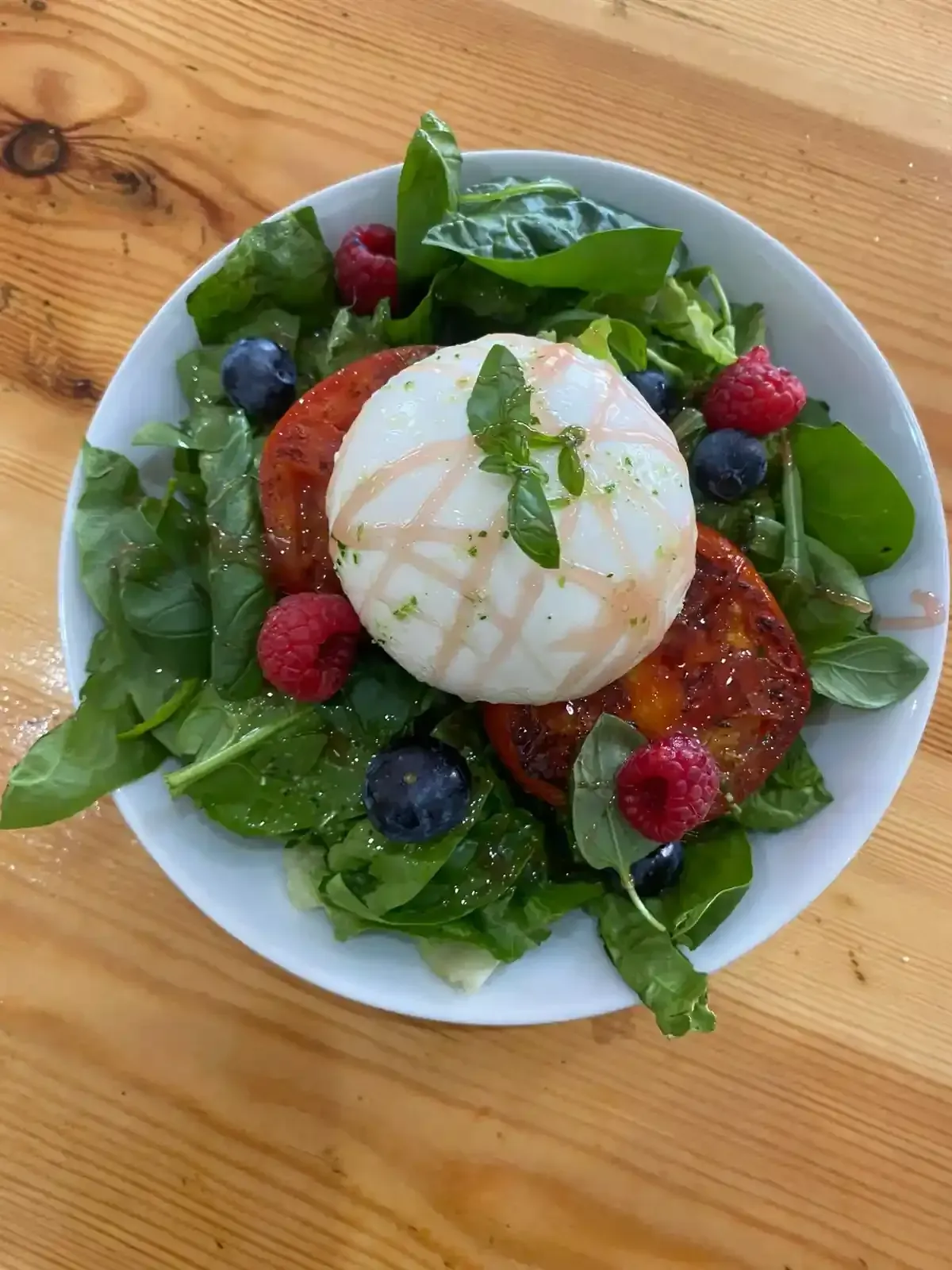 Ensalada con queso burrata, tomates fritos, bayas y espinacas en un plato blanco sobre una mesa de madera.