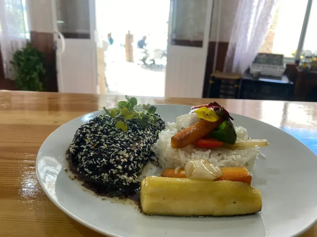 Plato con costra de sésamo, arroz, verduras y un trozo largo de pan amarillo. Colocado sobre una mesa de madera.