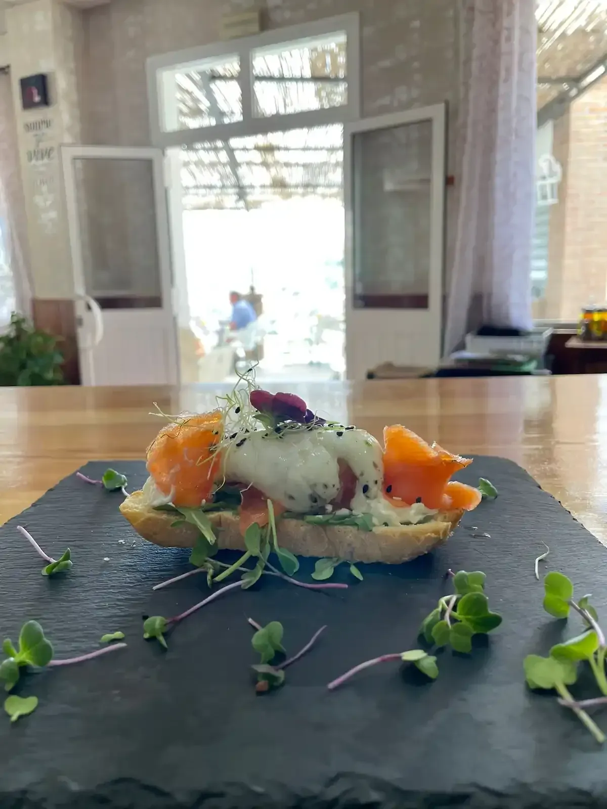 Aperitivo sobre pizarra negra: pan tostado con salmón ahumado, salsa cremosa y microgreens, con una puerta abierta al fondo.