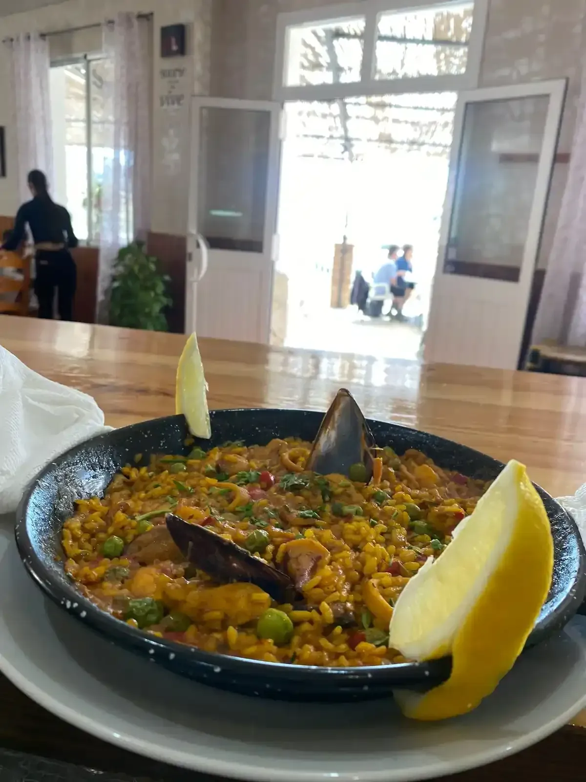 Paella en sartén negra con gajos de limón sobre una mesa, puerta al fondo.