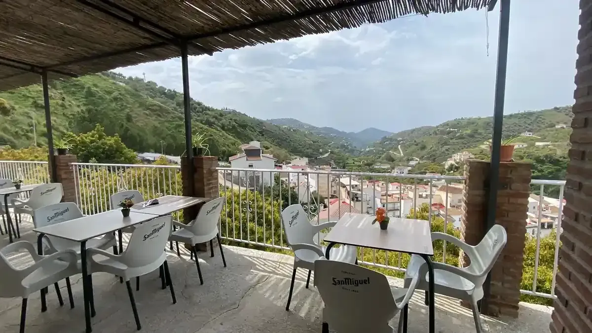 Restaurante al aire libre con mesas y sillas blancas, vistas a la montaña y al pueblo.
