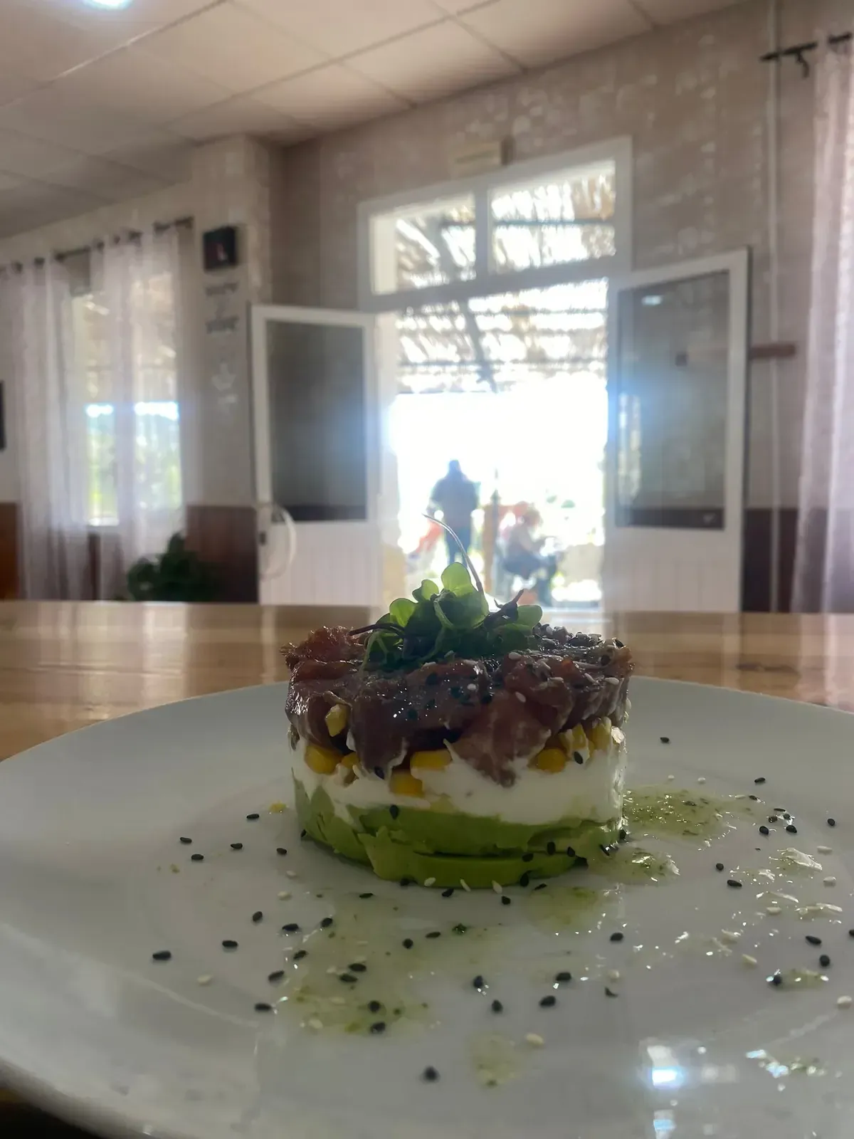 Plato gourmet apilado en un plato blanco; interior de edificio blanco con puertas abiertas en el fondo.