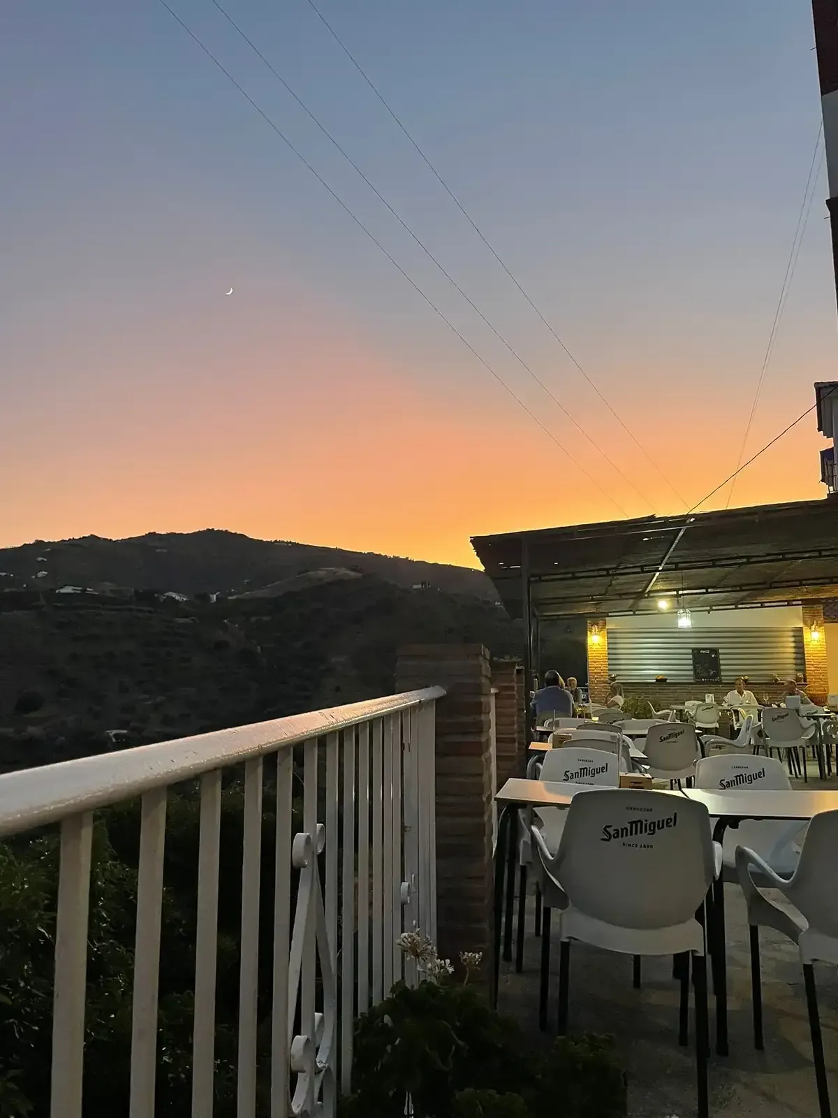Patio del restaurante al atardecer con vistas a las colinas. Mesas y sillas blancas, cielo naranja y azul.