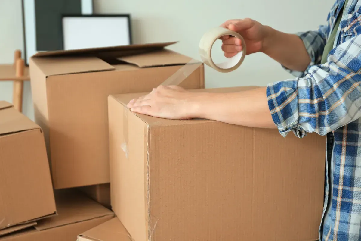 Persona colocando cinta adhesiva en una caja de cartón. Las cajas están apiladas en un interior