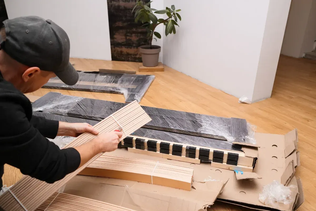 Persona desembalando piezas de muebles, sujetando un listón de madera, con otros componentes y embalajes en el suelo.