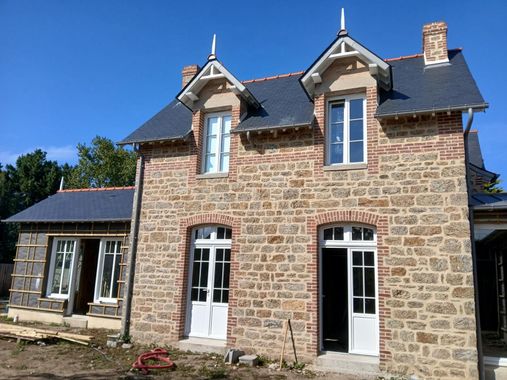 Une maison en pierre en construction, avec deux lucarnes à pignon, des portes et fenêtres à encadrement blanc et un toit de tuiles sombres.